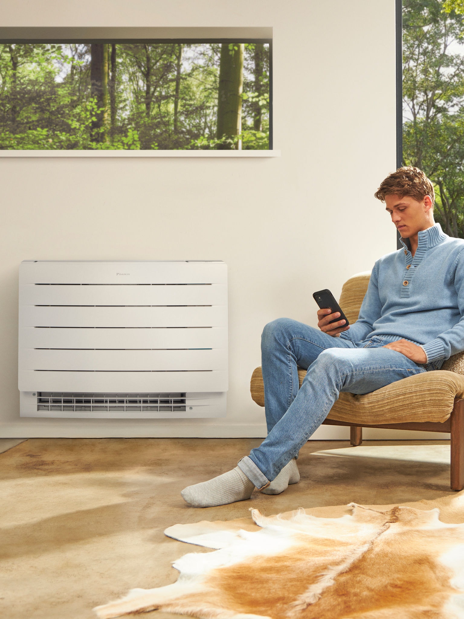 Moški sedi v svetli sobi in gleda v telefon s toplotno črpalko na steni za sabo