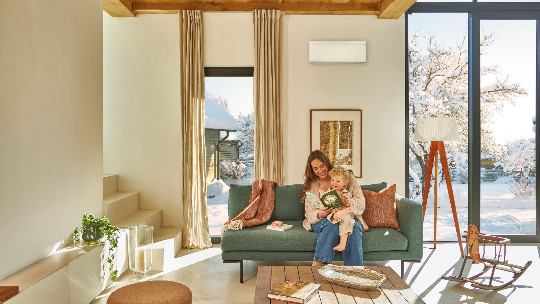 Mama sedi na kavču pod stensko toplotno črpalko zrak–zrak in bere otroško knjigo z malčkom v naročju, pri tem pa uživa v toploti, ko zunaj sneži